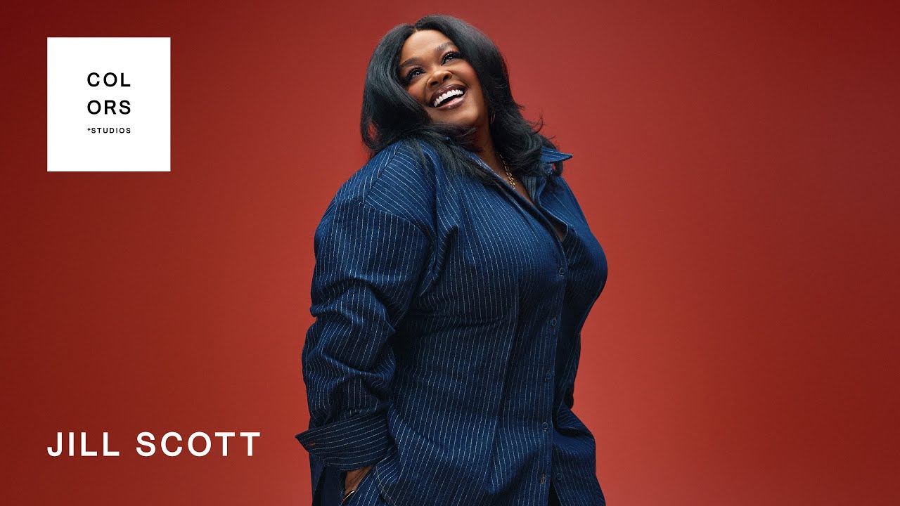 Jill Scott smiles in a blue pinstripe shirt against a red backdrop, COLORS Studios logo in the upper left.