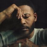 Moody close-up portrait of a man with a beard and short hair, eyes closed, head resting on his hand; Vevo logo appears in the bottom-left corner.