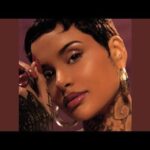 Close-up portrait of a woman with short dark hair, visible neck tattoos, and hoop earrings, wearing glossy makeup against a maroon background.