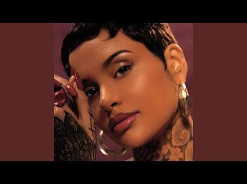 Close-up portrait of a woman with short dark hair, visible neck tattoos, and hoop earrings, wearing glossy makeup against a maroon background.