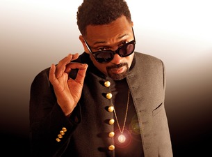 Portrait of a man in a black coat with gold buttons and sunglasses, posing with a hand near his face and a glowing pendant.
