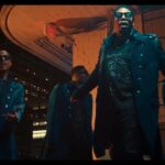 Group of four men in matching blue trench coats and sunglasses pose dramatically at night in a neon-lit urban setting, like a music video scene.