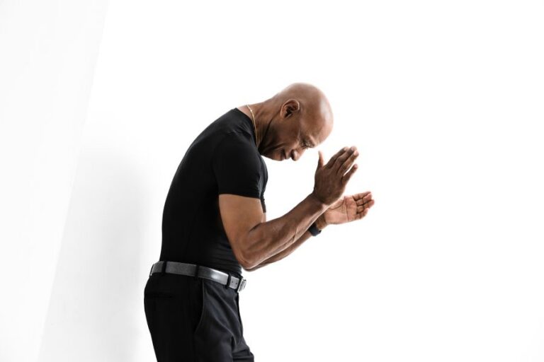 Bald man in a black athletic shirt bows with hands pressed together in a prayer-like gesture